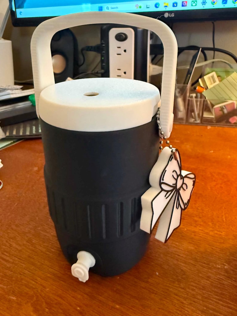 May include: A navy blue and white water cooler with a white handle and a white lid. The cooler has a spigot and a decorative bow-shaped charm. The cooler is designed to keep beverages cold.