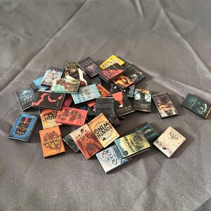May include: A collection of miniature paperback books with various titles and covers. The books are arranged in a pile on a gray fabric surface. Some of the titles include "The Glass Sword", "Throne of Glass", "A Court of Thorns and Roses", "The War of the Stars", "The Sieve and the Flame", "The Fourth Wing", "The Queen of Nothing", "Insurgent", "One", and "Land of Shadows".