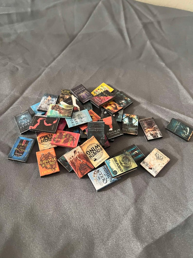 May include: A collection of miniature paperback books with various titles and covers. The books are arranged in a pile on a gray fabric surface. Some of the titles include "The Glass Sword", "Throne of Glass", "A Court of Thorns and Roses", "The War of the Stars", "The Sieve and the Flame", "The Fourth Wing", "The Queen of Nothing", "Insurgent", "One", and "Land of Shadows".