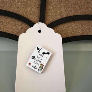 May include: A white tag with a scalloped edge and a small book attached to it. The book cover features a black and white illustration of a dragon, a sword, and flowers. The text on the book cover reads "ANY BOOK COVER HERE".