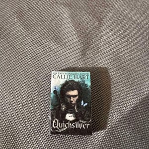 May include: A paperback book with a black cover and the title "Quicksilver" in white lettering. The author's name, Callie Hart, is printed in white on a black banner above the title. The cover features a man with dark hair and a serious expression, wearing a dark shirt and a white collar. There are two blue butterflies on the cover.