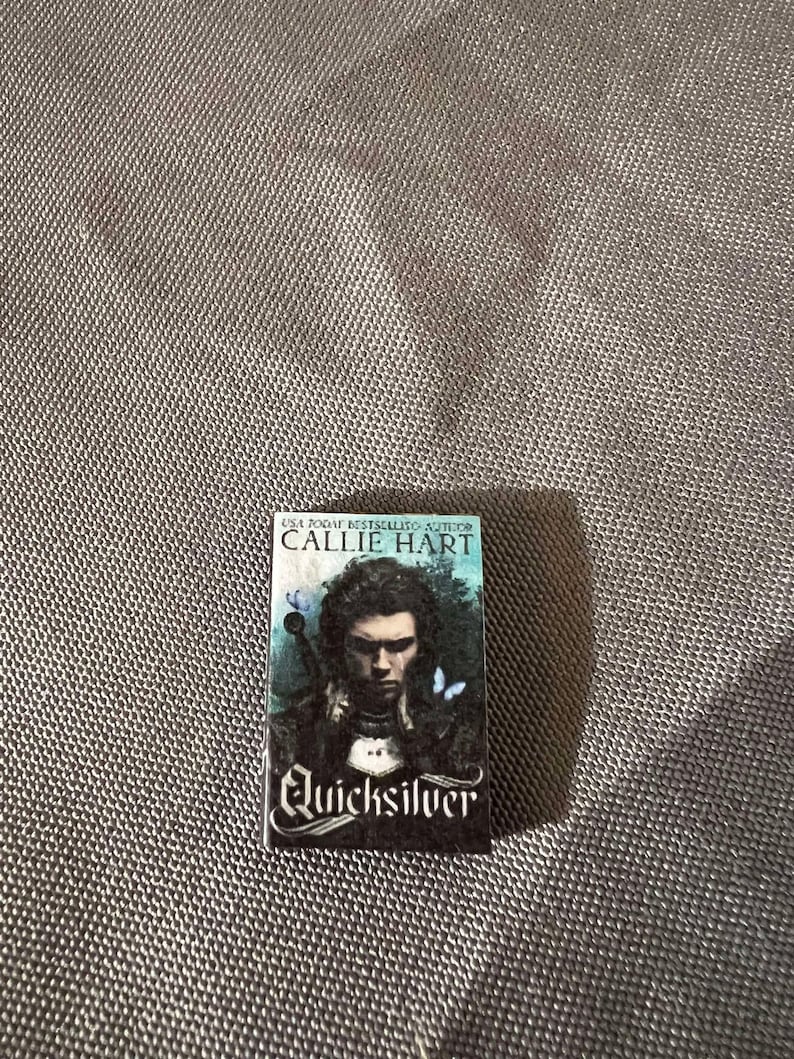 May include: A paperback book with a black cover and the title "Quicksilver" in white lettering. The author's name, Callie Hart, is printed in white on a black banner above the title. The cover features a man with dark hair and a serious expression, wearing a dark shirt and a white collar. There are two blue butterflies on the cover.