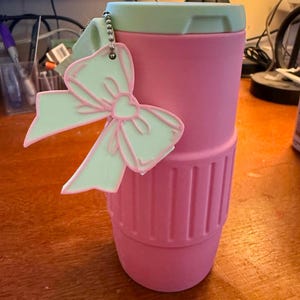 May include: A pink travel mug with a light green lid and a matching bow-shaped keychain. The mug has a ribbed design near the bottom. The bow keychain has a pink outline and a light green fill.