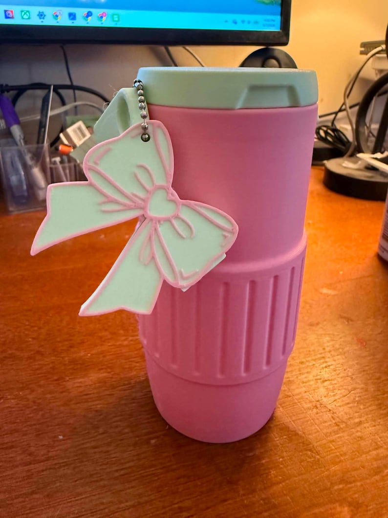 May include: A pink travel mug with a light green lid and a matching bow-shaped keychain. The mug has a ribbed design near the bottom. The bow keychain has a pink outline and a light green fill.