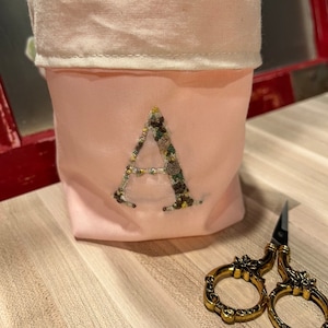 May include: A pink fabric pouch with a white top and an embroidered letter "A" in brown and green thread.