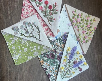 May include: A collection of handmade, triangular fabric bookmarks. Each bookmark features a different floral embroidery design, including roses, lavender, and daisies. The fabric backgrounds vary in color and pattern, with floral prints and solid colors.