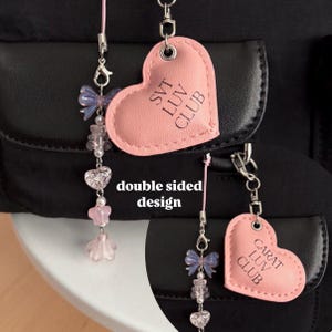 May include: Pink heart-shaped keychain with the text "SVT LUV CLUB" on one side and "CARAT LUV CLUB" on the other side. The keychain is attached to a black bag with a silver chain and a pink beaded charm.