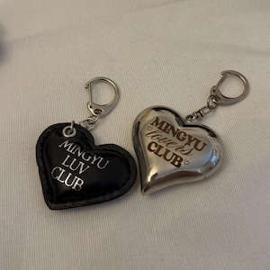 Pode incluir: Dois chaveiros em forma de coração. Um é preto com o texto "MINGYU LUV CLUB" em branco. O outro é prateado com o texto "MINGYU Lovers CLUB". Ambos têm argolas prateadas.