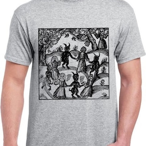 Op de afbeelding: Een zwart-witte illustratie van een groep mensen die in een cirkel dansen met een duivelse figuur in het midden. De illustratie speelt zich af in een bos met bomen en bloemen. De afbeelding is gedrukt op een grijs T-shirt.