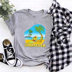 Puede incluir: Una camiseta gris con un gráfico de una escena de playa con palmeras y la palabra "WHATEVER" en amarillo. Zapatillas deportivas en blanco y negro y una camisa a cuadros están al lado de la camiseta.