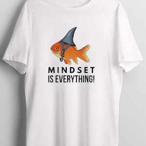 Puede incluir: Camiseta blanca con un gráfico de un pez dorado con un sombrero de aleta de tiburón. El texto "MINDSET IS EVERYTHING!" está impreso debajo del gráfico.