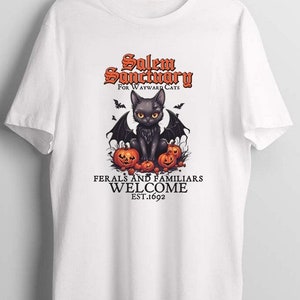 Może przedstawiać: Biała koszulka z czarnym kotem z skrzydłami i dyniami. Tekst głosi "Salem Sanctuary for Wayward Cats" i "Ferals and Familiars Welcome Est. 1692".