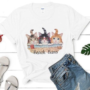 Pode incluir: Camiseta branca com um gráfico de três gatos de óculos de sol sentados numa praia com o texto "Beach Bums".