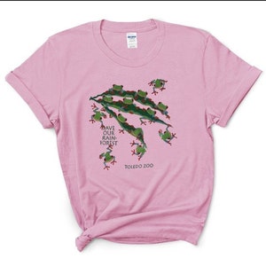 Op de afbeelding: Roze T-shirt met een groen kikkerontwerp en de tekst "Save Our Rain-Forest" en "Toledo Zoo".