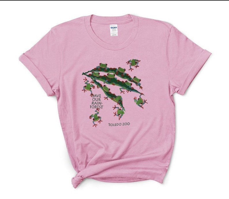 Op de afbeelding: Roze T-shirt met een groen kikkerontwerp en de tekst "Save Our Rain-Forest" en "Toledo Zoo".