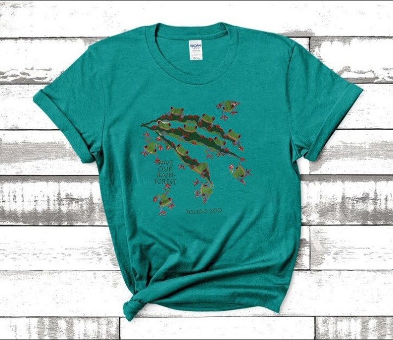 Op de afbeelding: Een turquoise t-shirt met een afbeelding van groene boomkikkers op een tak. De tekst "Save Our Rain Forest" is op het shirt gedrukt. Het shirt heeft een ronde hals en korte mouwen.