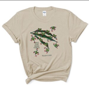 Op de afbeelding: Een beige T-shirt met een groen en rood kikkerontwerp. Het shirt heeft de tekst "Save Our Rain-Forest" en "Toledo Zoo" erop gedrukt.