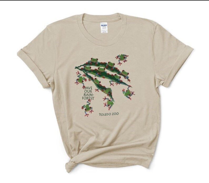 Op de afbeelding: Een beige T-shirt met een groen en rood kikkerontwerp. Het shirt heeft de tekst "Save Our Rain-Forest" en "Toledo Zoo" erop gedrukt.