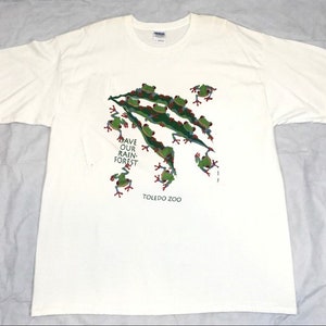 Op de afbeelding: Wit T-shirt met een groen, rood en blauw kikkerontwerp. De kikkers bevinden zich op een groen blad en de tekst "Save Our Rain Forest" en "Toledo Zoo" staat op het shirt gedrukt.