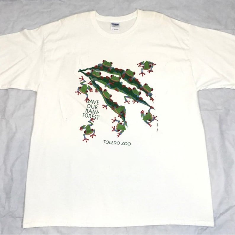 Op de afbeelding: Wit T-shirt met een groen, rood en blauw kikkerontwerp. De kikkers bevinden zich op een groen blad en de tekst "Save Our Rain Forest" en "Toledo Zoo" staat op het shirt gedrukt.