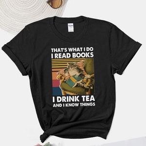 Könnte beinhalten: Schwarzes T-Shirt mit einer Grafik im Vintage-Stil, die eine Katze zeigt, die ein Buch liest und Tee trinkt. Der Text lautet "That's what I do I read books I drink tea and I know things."