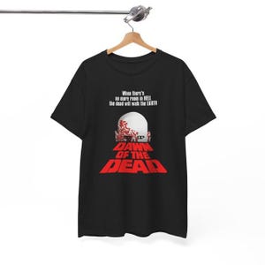Puede incluir: Camiseta negra con el texto "When there's no more room in HELL the dead will walk the EARTH" y el texto rojo "DAWN OF THE DEAD". El diseño incluye una cabeza de zombi.