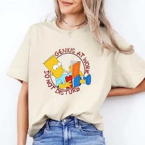 Op de afbeelding: Een beige t-shirt met een cartoon afbeelding van Bart Simpson die een boek leest en drinkt uit een rood bekertje. De tekst rond de afbeelding luidt "Genius at work, do not disturb".
