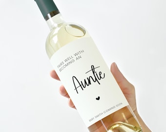 May include: A clear wine bottle with a white label that reads "Pairs well with becoming an Auntie" in black script. The bottle has a dark green cap and is filled with a light-colored liquid. A small black heart is below the word "Auntie".