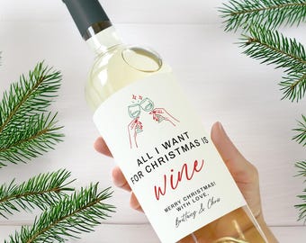 May include: A clear wine bottle with a dark green cap and a white label. The label reads "ALL I WANT FOR CHRISTMAS IS Wine" in red script, with a graphic of two wine glasses clinking. Green pine branches frame the bottle.