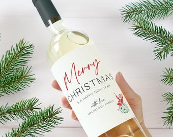 May include: A clear wine bottle with a dark green cap and a white label. The label reads "Merry Christmas & A Happy New Year" in red script. The bottle is held by a hand, with green pine branches in the background.