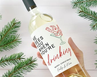 May include: A clear wine bottle with a dark green cap and a white label. The label features a red illustration of Santa Claus in a martini glass, with the text "He sees you when you're drinking" and "Merry Christmas!"