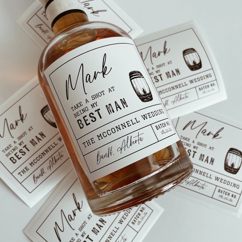 Op de afbeelding: Een wit etiket met zwarte tekst en een kleine afbeelding van een vat met de tekst "Mark Neem een ​​shot om te vieren dat je mijn getuige bent De McConnell Wedding Banff, Alberta Batch No. 08.10.24".