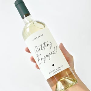 Könnte beinhalten: Eine klare Weinflasche mit grünem Verschluss und weißem Etikett. Auf dem Etikett steht "Cheers to Getting Engaged!" in eleganter Schrift, mit einem kleinen Herz und den Namen Michael & Kirstie sowie dem Datum 09.20.2026.