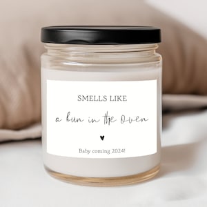 May include: A white candle with a black lid and a white label that says "Smells like a bun in the oven" with a small heart symbol and "Baby coming 2024!"