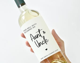May include: A clear wine bottle with a green cap and a white label. The label reads "PAIRS WELL WITH BECOMING AN Aunt & Uncle" with a heart symbol and "BABY SMITH COMING SOON". The bottle is filled with a light-colored liquid.