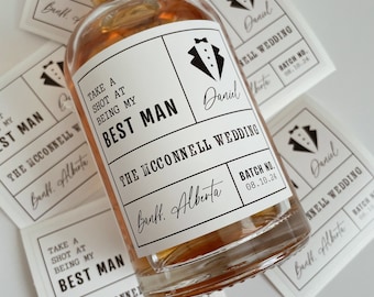 May include: A clear glass bottle with a white label that says "Take a shot at being my Best Man" and "The McConnell Wedding" with a black tuxedo graphic. The label also includes the text "Banff, Alberta" and "Batch No. 08.10.24".
