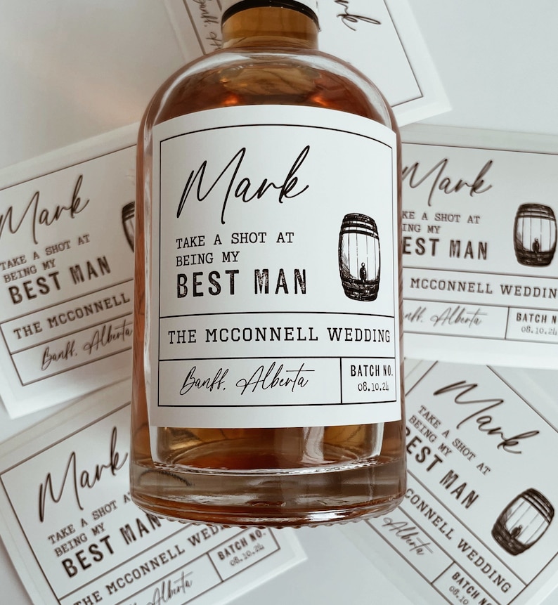 Op de afbeelding: Een bruine glazen fles met een wit etiket waarop staat "Mark Take a Shot at Being My Best Man The McConnell Wedding Banff, Alberta Batch No. 08.10.21". Het etiket bevat ook een kleine afbeelding van een vat.