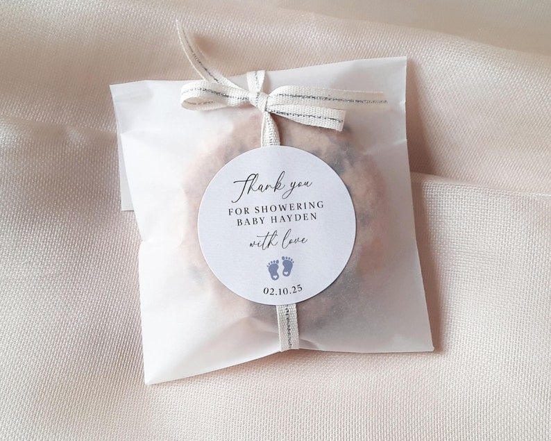 May include: White favour bag with a round thank you sticker. The sticker says "Thank you for showering baby Hayden with love" and has a date of 02.10.25 and two blue footprints.
