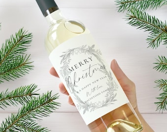 May include: A clear wine bottle with a dark green cap and a label that reads "Merry Christmas" in elegant script. The label is decorated with a wreath design. The bottle is held in front of a white background with green pine branches.