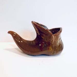 May include: A brown ceramic shoe-shaped planter with a glossy finish. The planter has a curved toe, a tall, open top, and a textured appearance. The color varies from light to dark brown, with some areas appearing almost black.