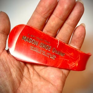 May include: A red shoehorn with white marbling, held in a hand. The shoehorn has the text "MASON SHOE Dealer" and "MASON SHOE MFG. CO. CHIPPEWA FALLS, WIS." in silver lettering. The text "A GIFT FROM YOUR" is above the main text.