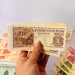 May include: A stack of vintage U.S. Department of Agriculture Food Coupons. The coupons are beige with brown ink, featuring illustrations of the Declaration of Independence and the Liberty Bell. Each coupon is worth one dollar.