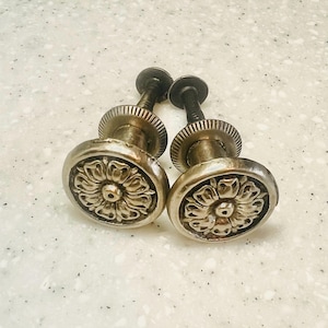 May include: Two antique silver-toned drawer pulls. Each pull features a detailed floral design on the face and a threaded stem. The pulls are likely made of metal and are designed for furniture restoration or decorative use.