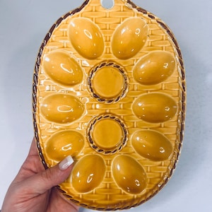 May include: A yellow ceramic egg serving dish with a woven basket design. The dish has 10 egg-shaped indentations and two circular indentations. The rim is brown. The dish is held by a person.