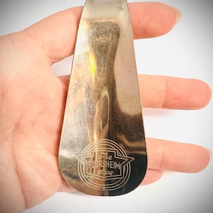 May include: A vintage silver-toned shoe horn with a rounded handle and a flared end. The shoe horn has the text "The Florsheim Shoe" in a circular emblem. The item is held in a hand against a white background.