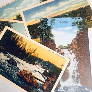 May include: A collection of vintage postcards featuring scenic landscapes. The postcards depict waterfalls, lakes, and forests with blue skies and colorful sunsets. Text on the postcards includes "Dead River, Marquette, Mich." and "Black River Falls, Near Ishpeming, Michigan - 14."