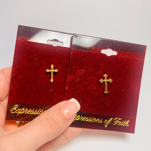 May include: Two gold-colored cross earrings on red velvet cards. The cards have the text "Expressions of Faith" in gold lettering. The earrings are displayed on a white background.