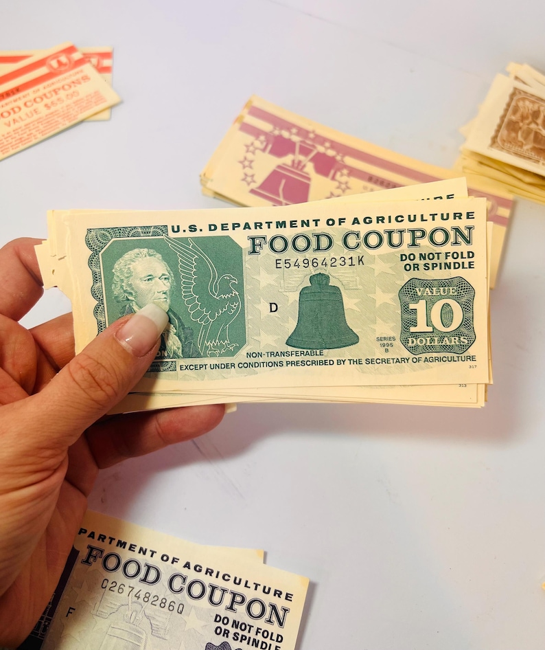 May include: A stack of U.S. Department of Agriculture Food Coupons, with a $10 value visible. The coupons feature an image of the Liberty Bell and the text "Food Coupon". Other coupons with different values are also present.
