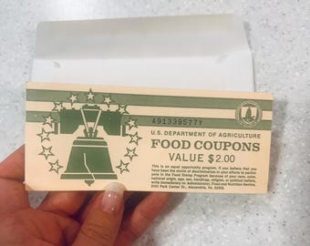 May include: A vintage U.S. Department of Agriculture food coupon, featuring a green Liberty Bell design surrounded by stars. The coupon, with a value of $2.00, includes text about equal opportunity and the Food Stamp Program. The coupon is held in a white envelope.