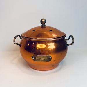 May include: A copper potpourri pot with a lid and two handles. The pot has a round shape and a shiny finish. The lid has small holes and a small knob on top. The word "POTPOURRI" is on a small plaque on the front.